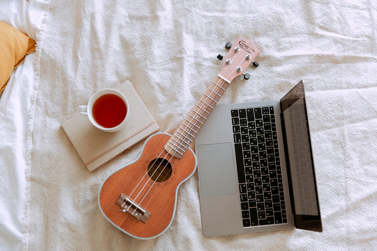 who-we-are A cozy composition featuring a ukulele, laptop, and cup of tea on a bed for a relaxing indoor ambiance.
